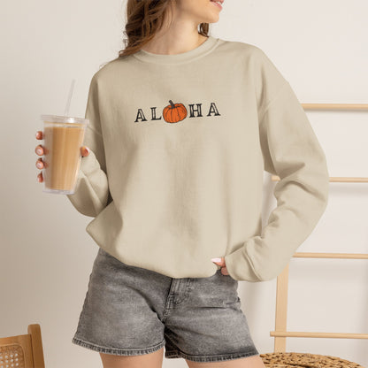 A woman wearing a beige crewneck sweatshirt with 'ALOHA' printed across the chest, where the 'O' is replaced by an orange pumpkin graphic. She is holding a clear plastic iced coffee cup and wearing gray denim shorts in a brightly lit indoor setting.