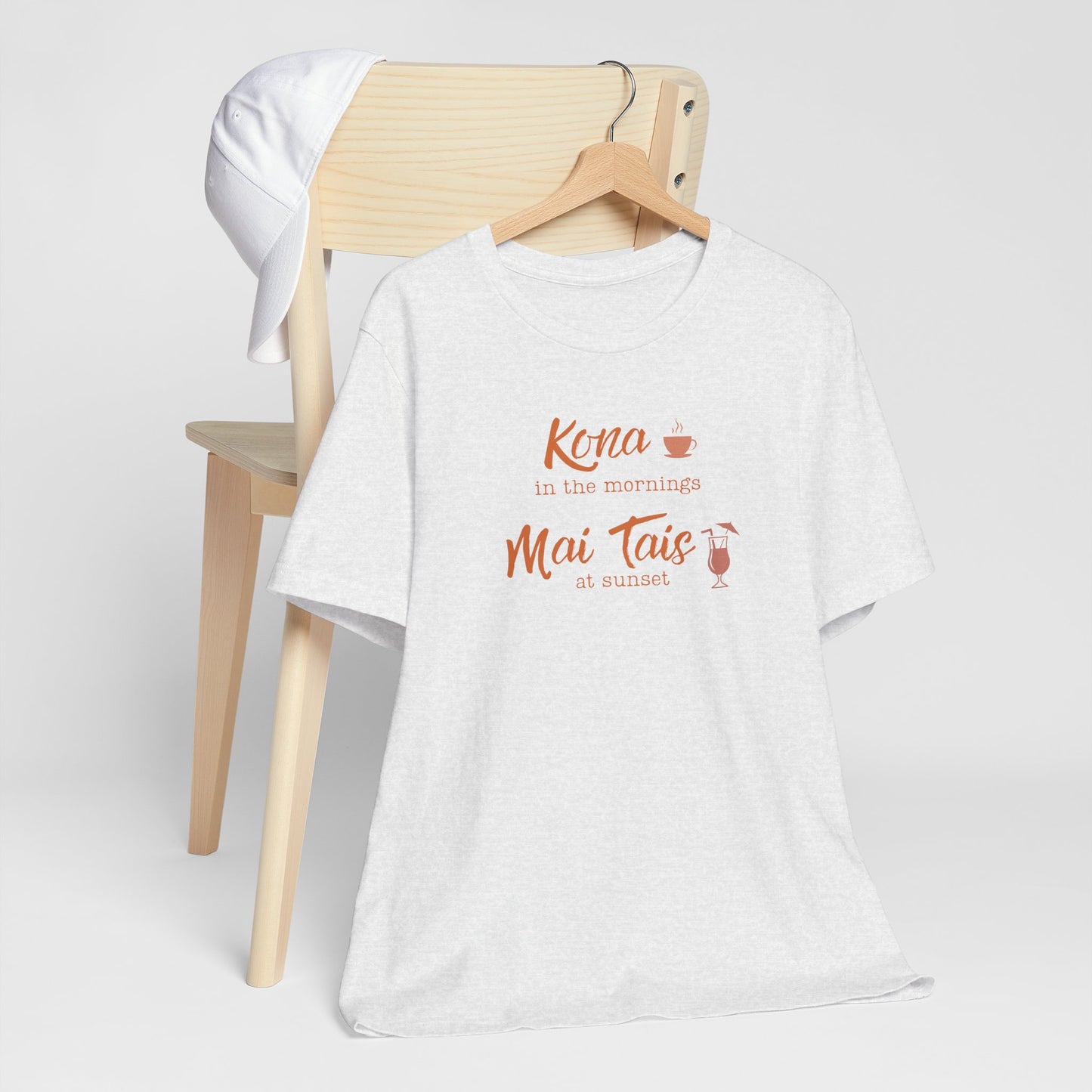 A white heather graphic t-shirt hanging on a wooden hanger, styled in front of a light wood chair with a white towel draped over the backrest. The shirt features a coral orange graphic that reads 'Kona in the mornings Mai Tais at sunset' with small coffee and cocktail illustrations.
