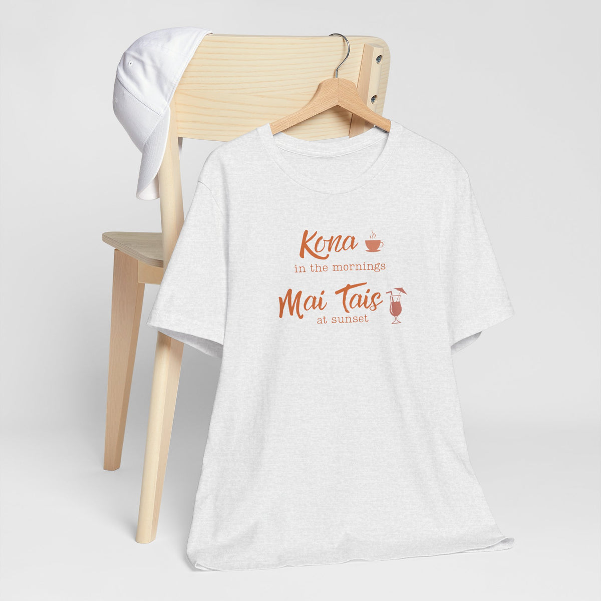 A white heather graphic t-shirt hanging on a wooden hanger, styled in front of a light wood chair with a white towel draped over the backrest. The shirt features a coral orange graphic that reads 'Kona in the mornings Mai Tais at sunset' with small coffee and cocktail illustrations.