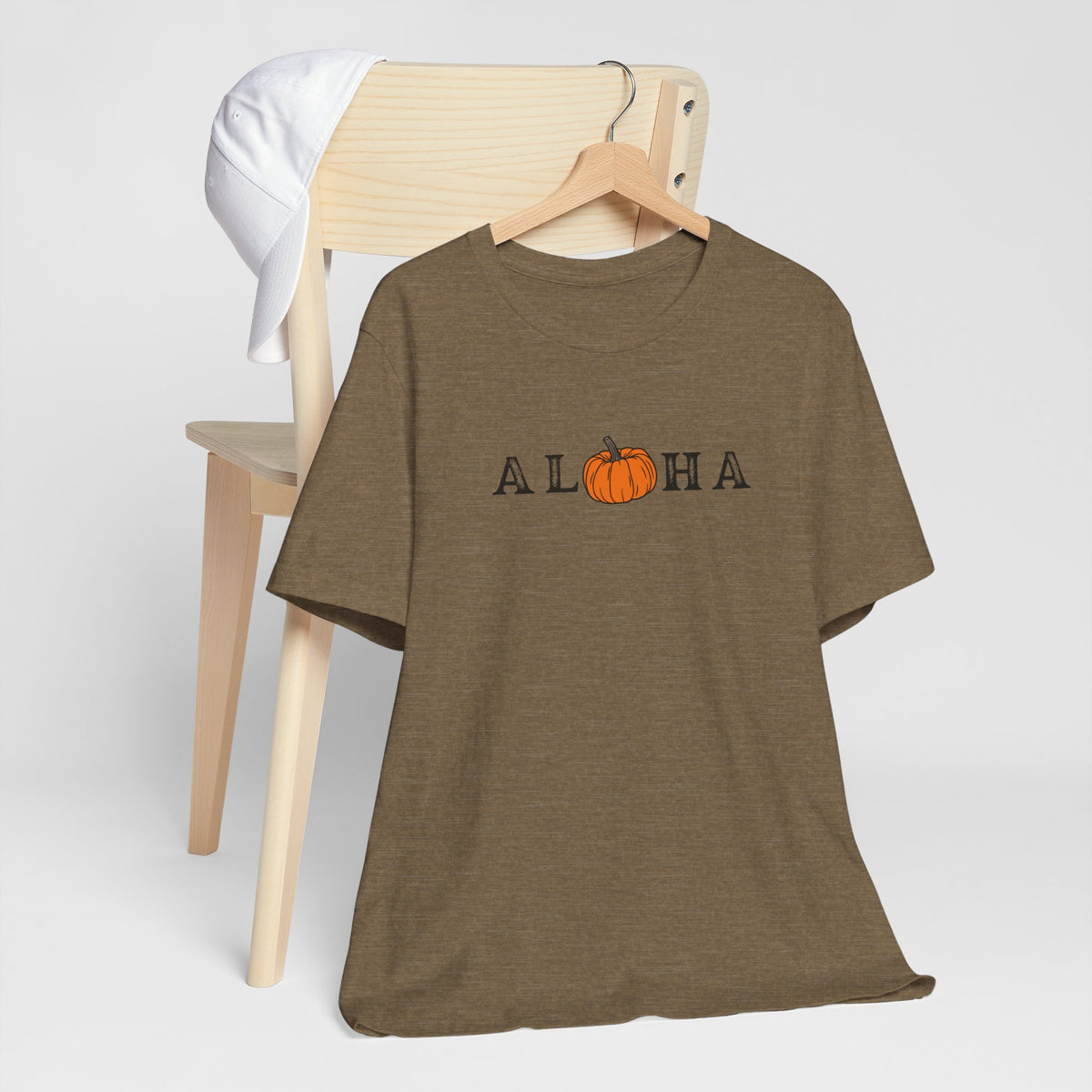 A heather olive short-sleeve t-shirt on a wooden hanger, featuring the word 'ALOHA' with a pumpkin graphic replacing the 'O'. A light wood chair and a white towel are in the background against a light gray studio backdrop.