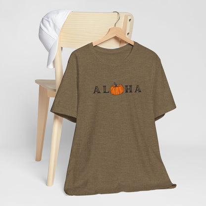 A heather olive short-sleeve t-shirt on a wooden hanger, featuring the word 'ALOHA' with a pumpkin graphic replacing the 'O'. A light wood chair and a white towel are in the background against a light gray studio backdrop.
