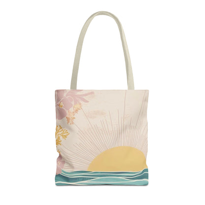 A canvas tote bag with a minimalist tropical sunset print, featuring a pale yellow sun, teal ocean waves, and pink hibiscus flowers on a cream background, with light beige shoulder straps.
