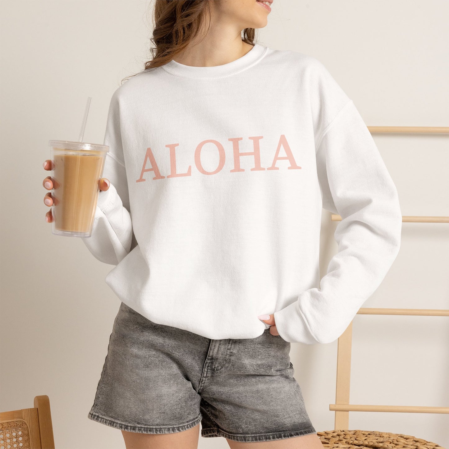 A woman wearing an oversized crewneck sweatshirt in white with the word 'ALOHA' printed across the chest in a light pink color. She is holding a clear plastic cup of iced coffee and wearing gray denim shorts, set against a warm, neutral indoor background.