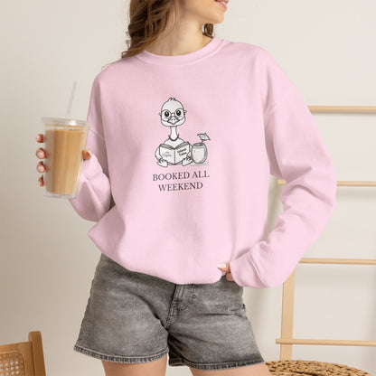 A woman wearing a light pink crewneck sweatshirt with a cartoon duck graphic and the text 'BOOKED ALL WEEKEND' printed on the front. She is holding an iced coffee and wearing gray denim shorts in a brightly lit indoor setting.