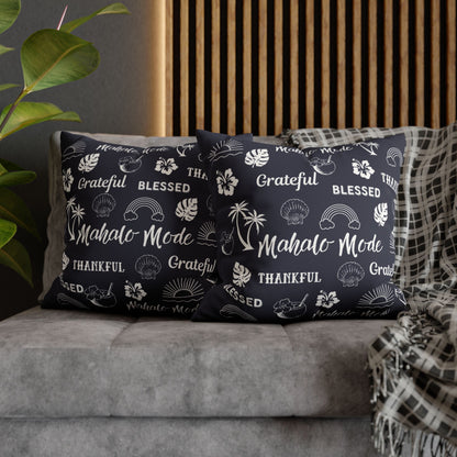 Two navy blue throw pillows with white tropical-themed text and illustrations, styled on a gray sofa with a plaid throw blanket and a wooden slat wall background.