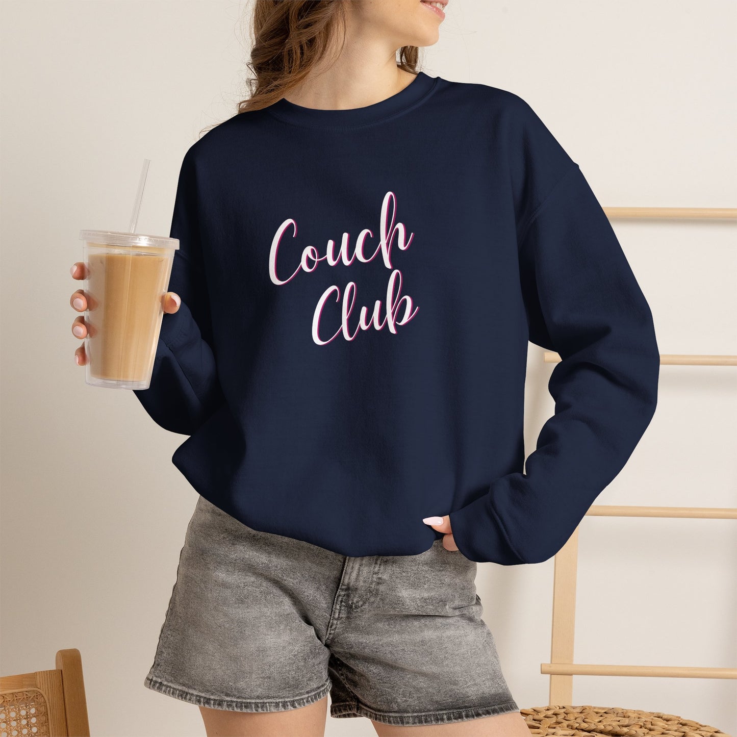 A woman wearing a navy blue crewneck sweatshirt with 'Couch Club' printed in pink script across the chest, paired with gray denim shorts and holding an iced coffee.