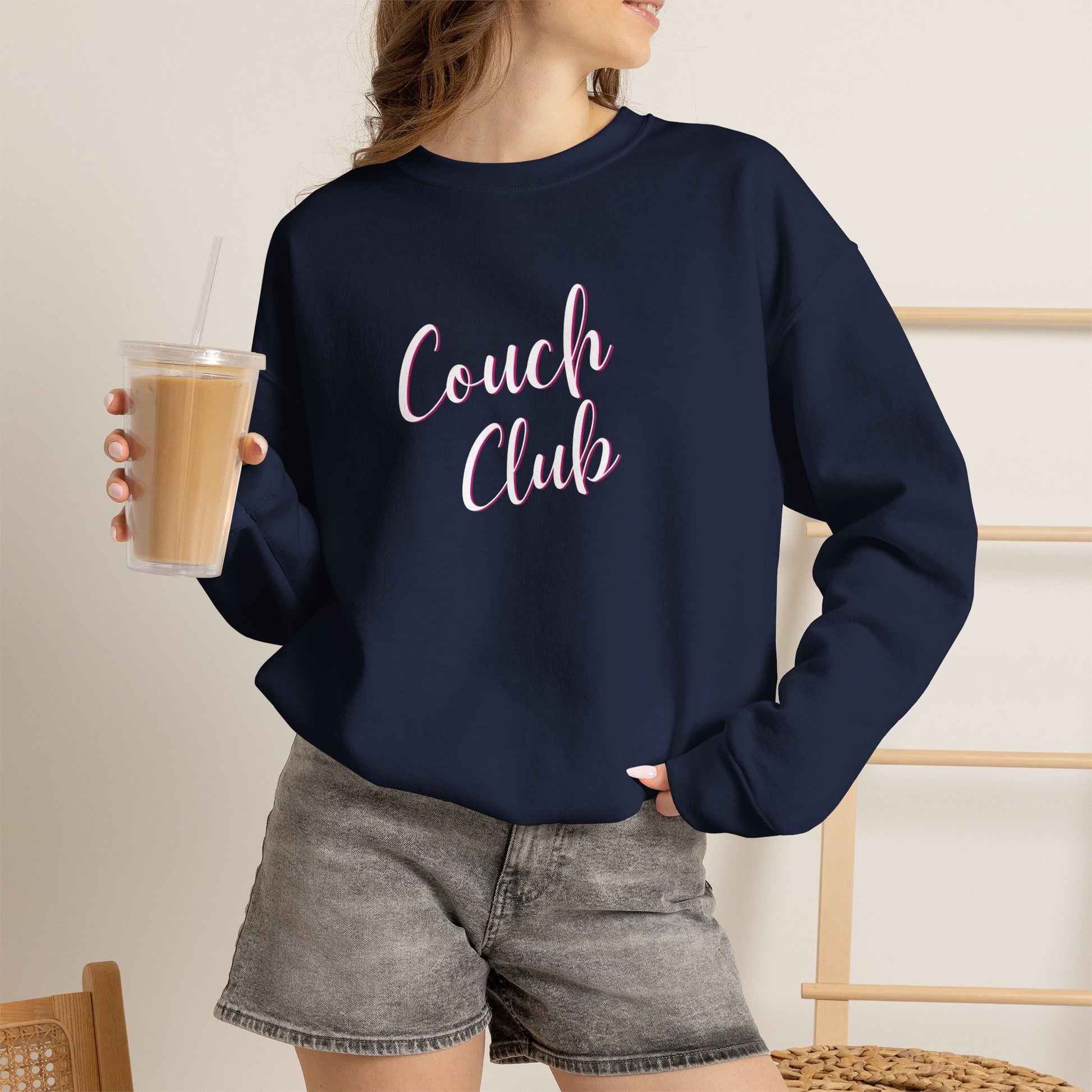 A woman wearing a navy blue crewneck sweatshirt with 'Couch Club' printed in pink script across the chest, paired with gray denim shorts and holding an iced coffee.