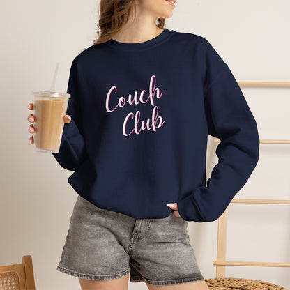 A woman wearing a navy blue crewneck sweatshirt with 'Couch Club' printed in pink script across the chest, paired with gray denim shorts and holding an iced coffee.