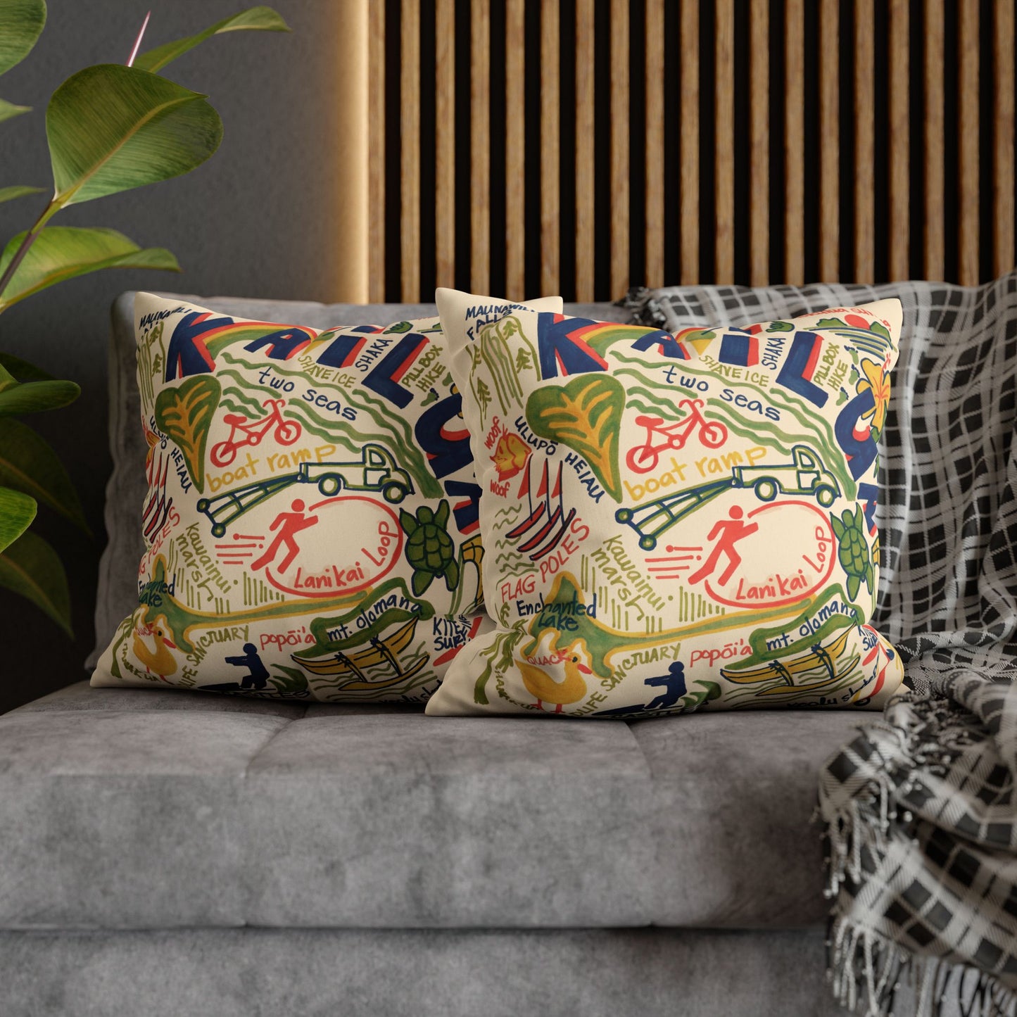 Two decorative throw pillows with a colorful vintage-style map illustration pattern of Kailua Oahu, styled on a gray sofa next to a black-and-white plaid throw blanket.