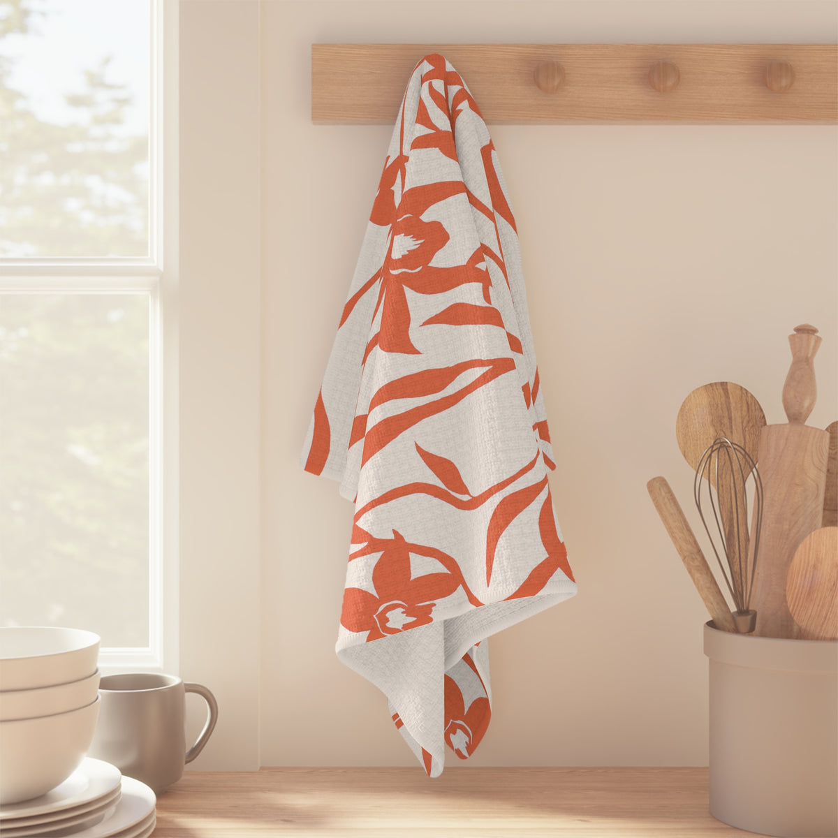 A rectangular white kitchen towel or tea towel featuring a bold coral orange floral pattern of orchids and leaves, shown hanging off of wood peg in a kitchen setting.