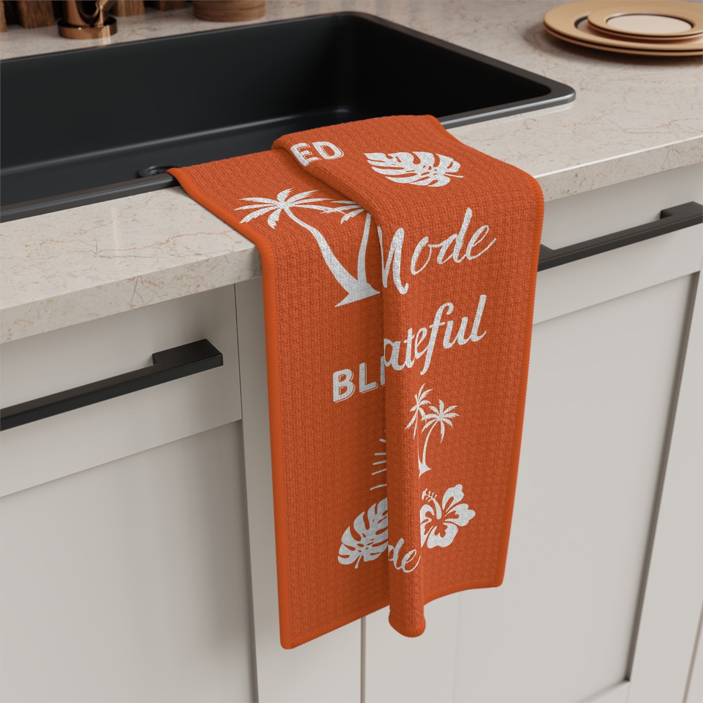 An orange waffle-weave kitchen towel with white tropical and text graphics hangs over the edge of a black sink in a modern kitchen setting.