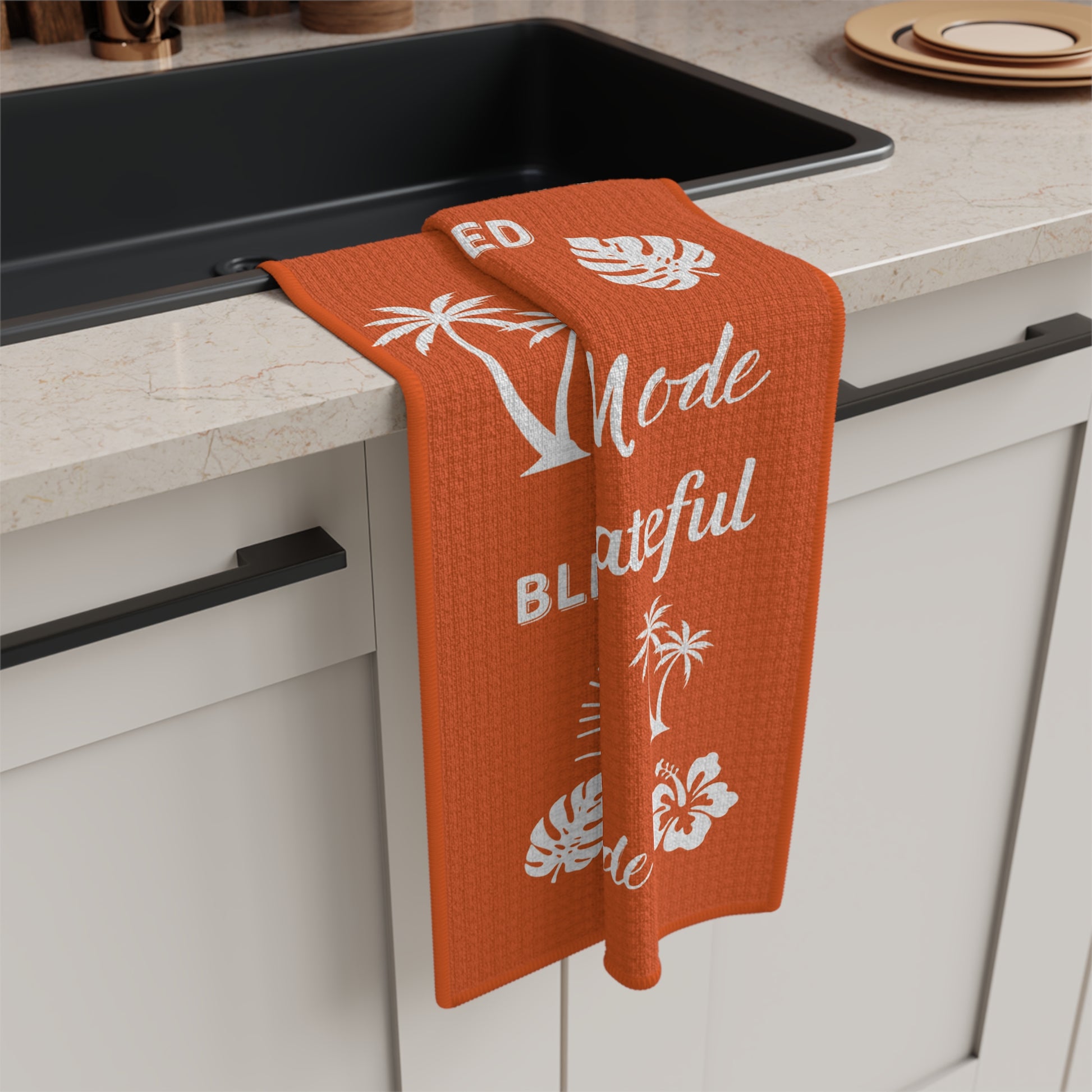 An orange waffle-weave kitchen towel with white tropical and text graphics hangs over the edge of a black sink in a modern kitchen setting.