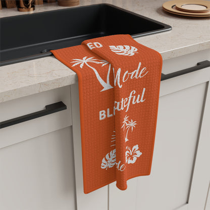 An orange waffle-weave kitchen towel with white tropical and text graphics hangs over the edge of a black sink in a modern kitchen setting.