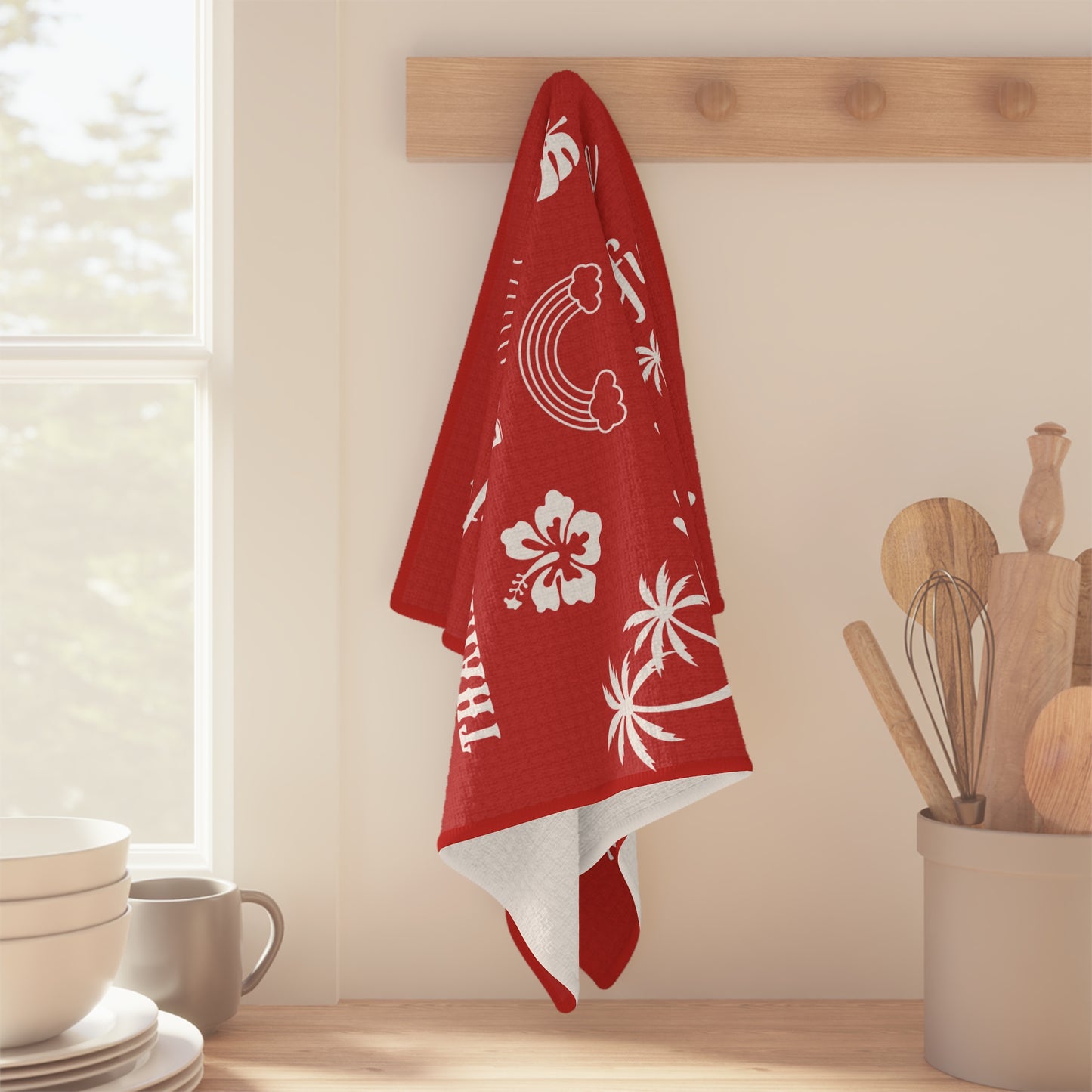 A red kitchen towel with white tropical and Hawaiian-themed prints hangs from a wooden peg on a wall in a bright kitchen setting.