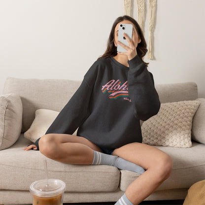A woman sits cross-legged on a beige couch taking a mirror selfie while wearing a dark charcoal crewneck sweatshirt with a colorful retro 'Aloha' graphic. She is wearing gray striped socks and holding an iced coffee in the foreground.