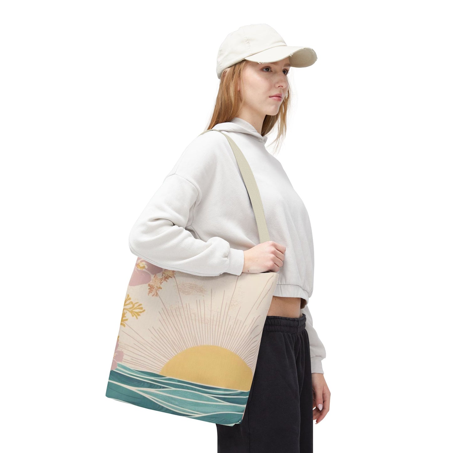 A woman wearing a white cap, white cropped hoodie, and black sweatpants carries a canvas tote bag with a sunset and ocean design. The bag has a beige base with a yellow sun, pink clouds, and teal waves, shown against a plain white background.