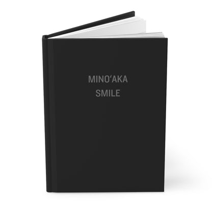 A hardcover notebook or journal standing upright on a white background. The cover is matte black with the words 'MINO'AKA SMILE' printed in a light gray serif font near the top.
