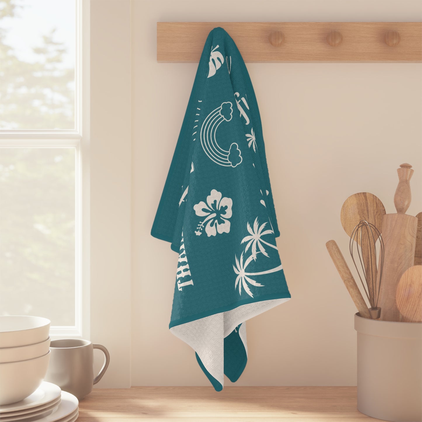 A teal kitchen towel with white tropical and beach-themed prints hangs from a wooden peg on a kitchen wall. The towel features white illustrations of palm trees, a sun, a hibiscus flower, and the word 'mahalo mode'.