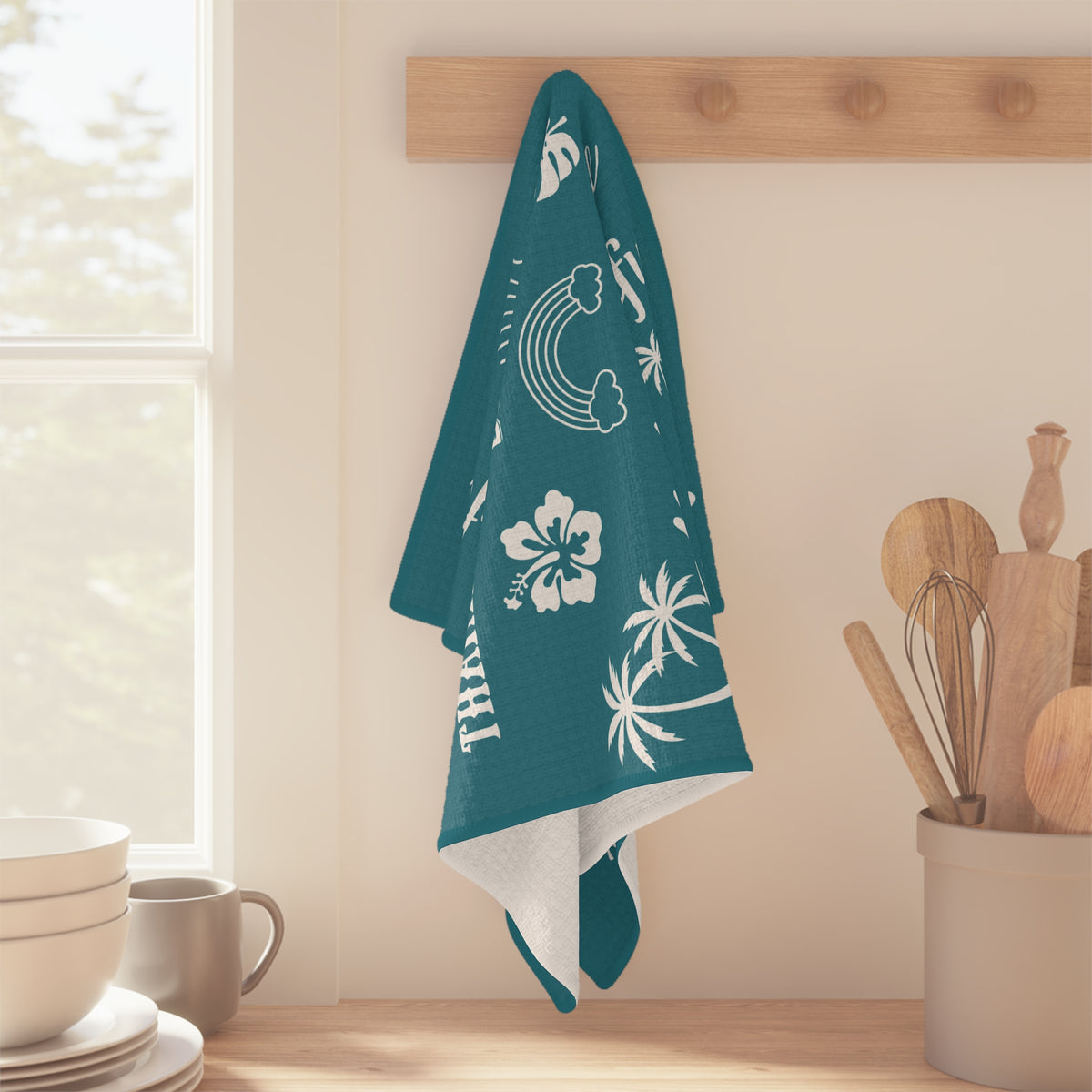 A teal kitchen towel with white tropical and beach-themed prints hangs from a wooden peg on a kitchen wall. The towel features white illustrations of palm trees, a sun, a hibiscus flower, and the word 'mahalo mode'.