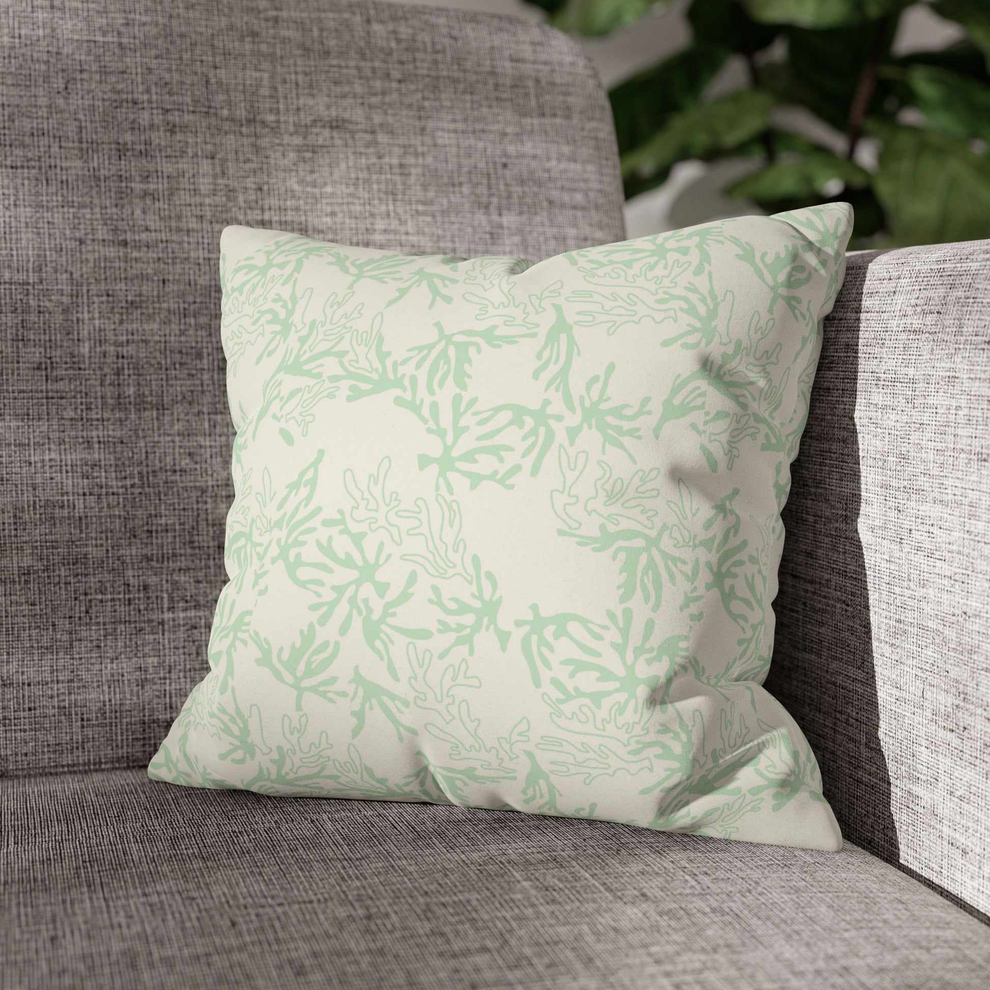 A square throw pillow featuring a light sage green coral branch pattern on an off-white background, on a grey couch with a plant behind the couch