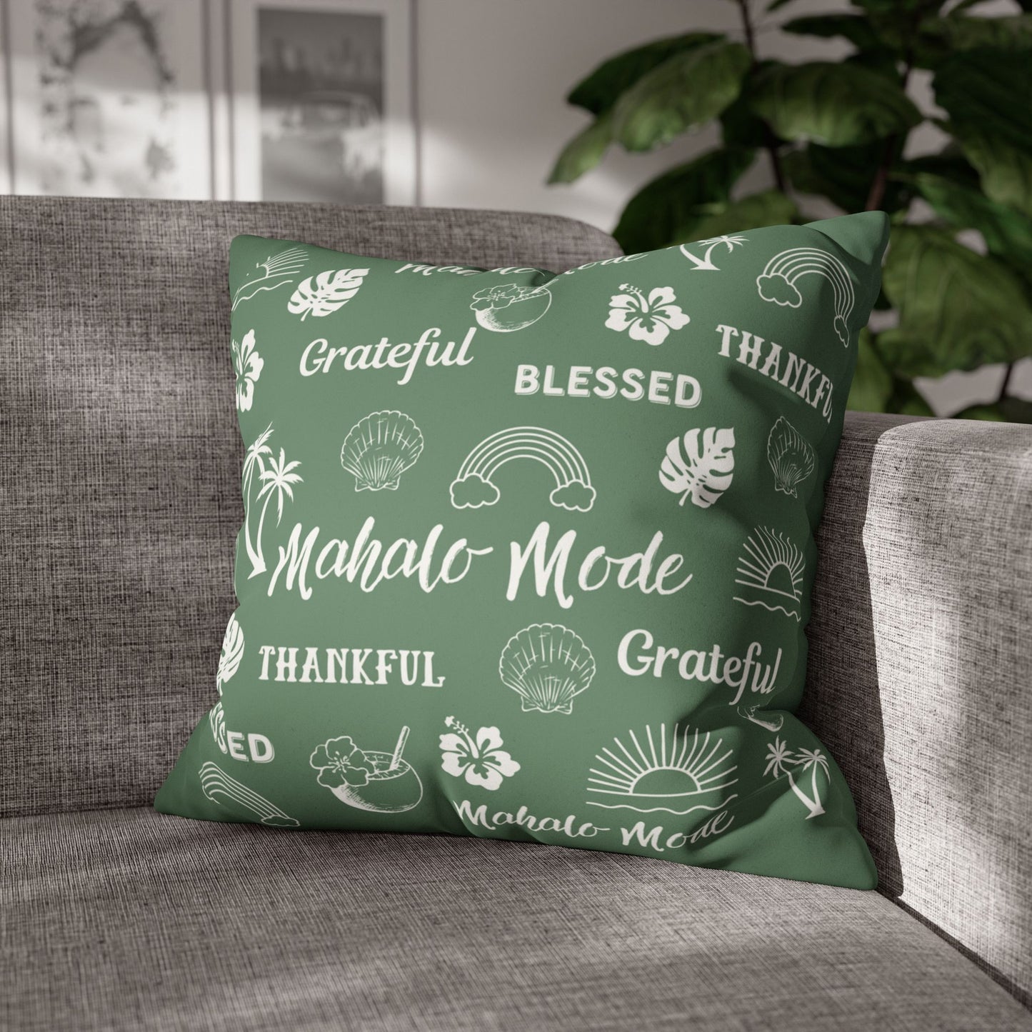 A square throw pillow in sage green with a white tropical-themed print featuring the words 'Grateful', 'Blessed', 'Thankful', and 'Mahalo Mode' along with icons like rainbows, hibiscus flowers, and seashells, resting on a gray fabric sofa with a blurred indoor background.