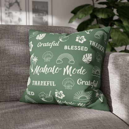 A square throw pillow in sage green with a white tropical-themed print featuring the words 'Grateful', 'Blessed', 'Thankful', and 'Mahalo Mode' along with icons like rainbows, hibiscus flowers, and seashells, resting on a gray fabric sofa with a blurred indoor background.
