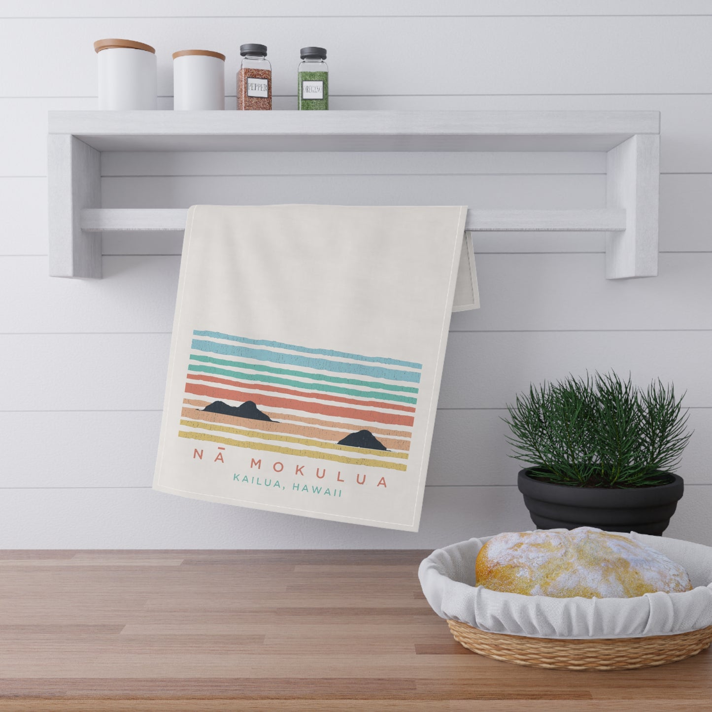 A cream-colored kitchen towel with a colorful retro sunset graphic and the text 'NÁ MOKULUA KAILUA, HAWAII' hanging from a white wall rack. The scene is styled in a bright kitchen with a wooden floor, a shelf with jars, a potted plant, and a basket with bread.