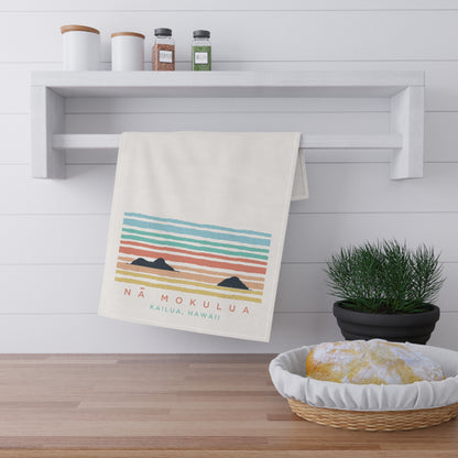A cream-colored kitchen towel with a colorful retro sunset graphic and the text 'NÁ MOKULUA KAILUA, HAWAII' hanging from a white wall rack. The scene is styled in a bright kitchen with a wooden floor, a shelf with jars, a potted plant, and a basket with bread.
