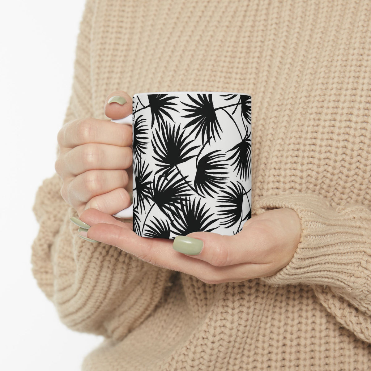 A person in a beige knit sweater holds a white ceramic mug with a black palm leaf pattern against a clean white background.
