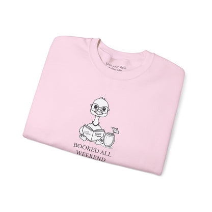 A folded light pink crewneck t-shirt featuring a cartoon nene goose character reading a book with the text 'BOOKED ALL WEEKEND' underneath, displayed on a clean white background.