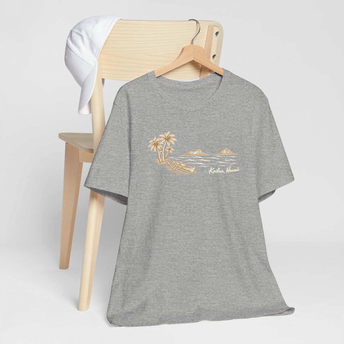 Athletic Heather unisex graphic tee with hand-drawn Kailua canoe, palm trees and Mokulua Islands artwork hanging from a wood chair