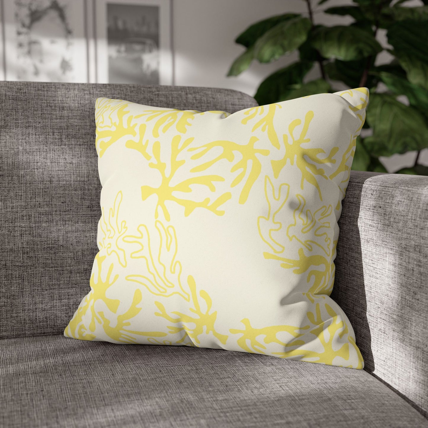 A square throw pillow with a yellow coral-like abstract pattern on an off-white background, resting on a gray textured sofa with a green plant in the background.