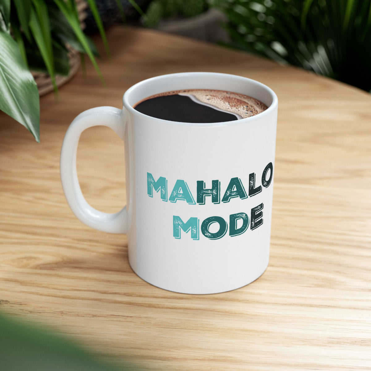 A white ceramic coffee mug with the words 'MAHALO MODE' printed in teal and dark gray lettering, filled with black coffee and sitting on a light wood table with green plants in the background.