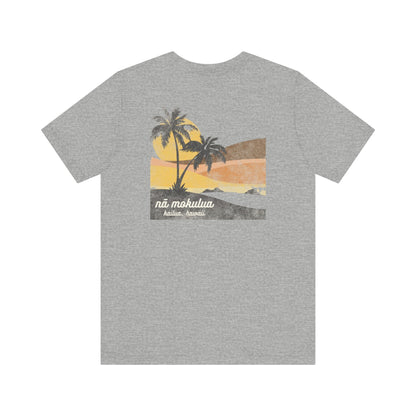 A heather gray short-sleeve t-shirt with a graphic print of a tropical sunset scene featuring palm trees, a yellow sun, and the text 'ná mokulua' and 'Kailua, Hawaii' on the back.