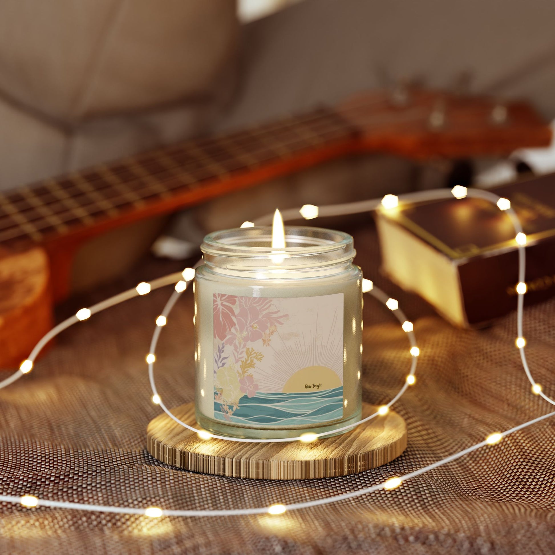 A lit glass jar candle with a tropical ocean sunset label sits on a wooden coaster, surrounded by warm white fairy lights, with a blurred guitar and book in the background.