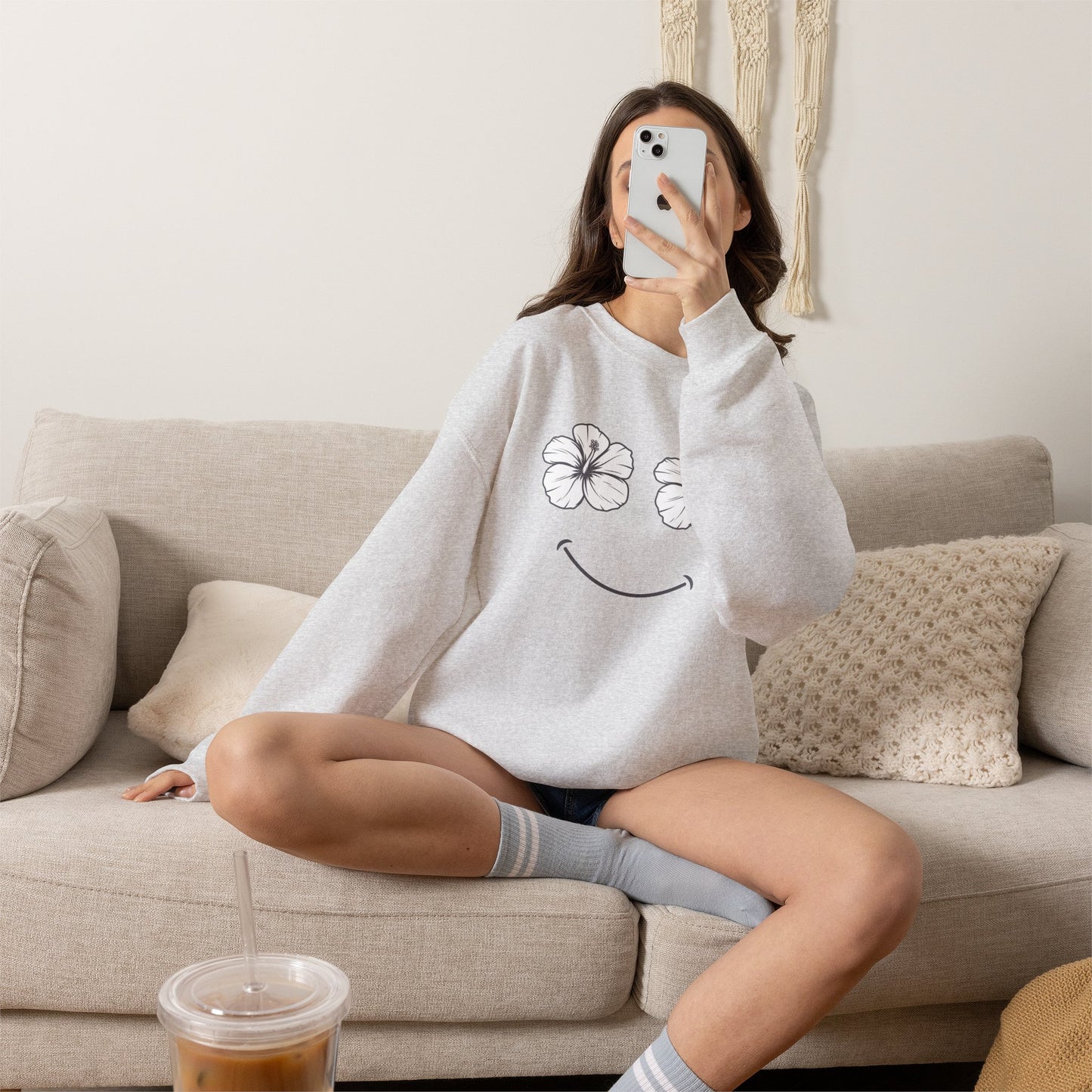 Person wearing a light gray sweatshirt sitting on a beige couch with a cup of coffee on a table wearing a Happy Hibiscus Sweatshirt featuring a cheerful hibiscus flower smile design inspired by island life and aloha style.