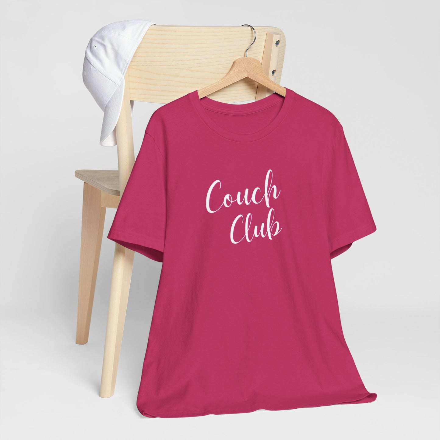 A dark pink short-sleeve t-shirt on a wooden hanger, styled in front of a light wood chair with a white cap. The t-shirt features the text 'Couch Club' in white script.