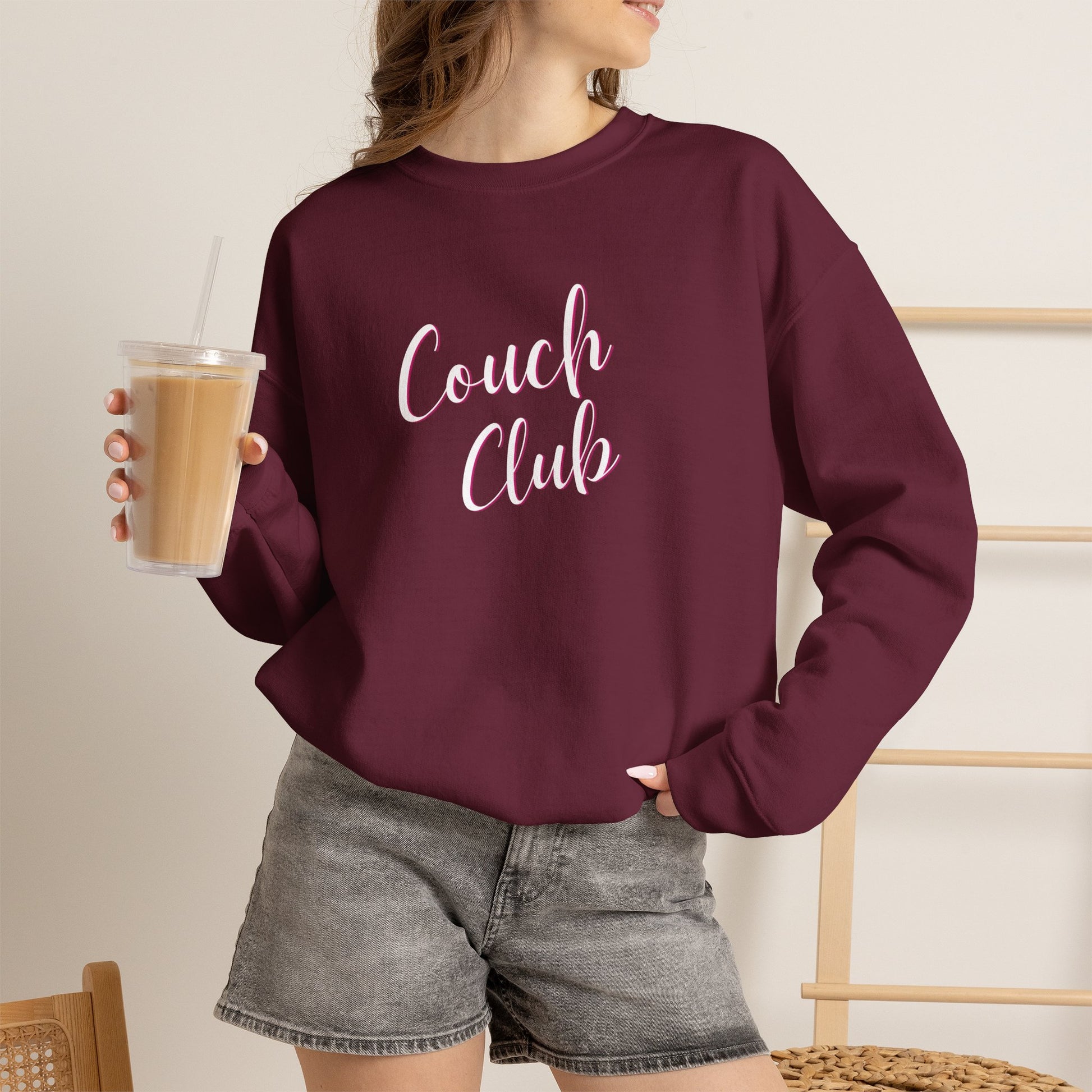 A woman wearing a maroon crewneck sweatshirt with 'Couch Club' printed in white script across the chest, holding an iced coffee in a bright indoor setting.