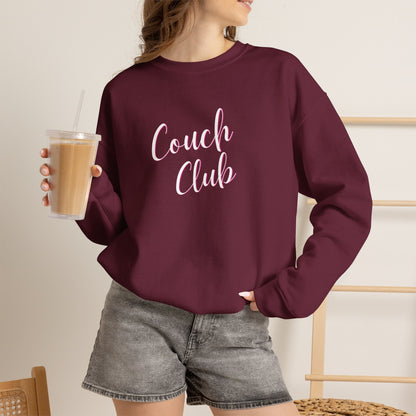 A woman wearing a maroon crewneck sweatshirt with 'Couch Club' printed in white script across the chest, holding an iced coffee in a bright indoor setting.