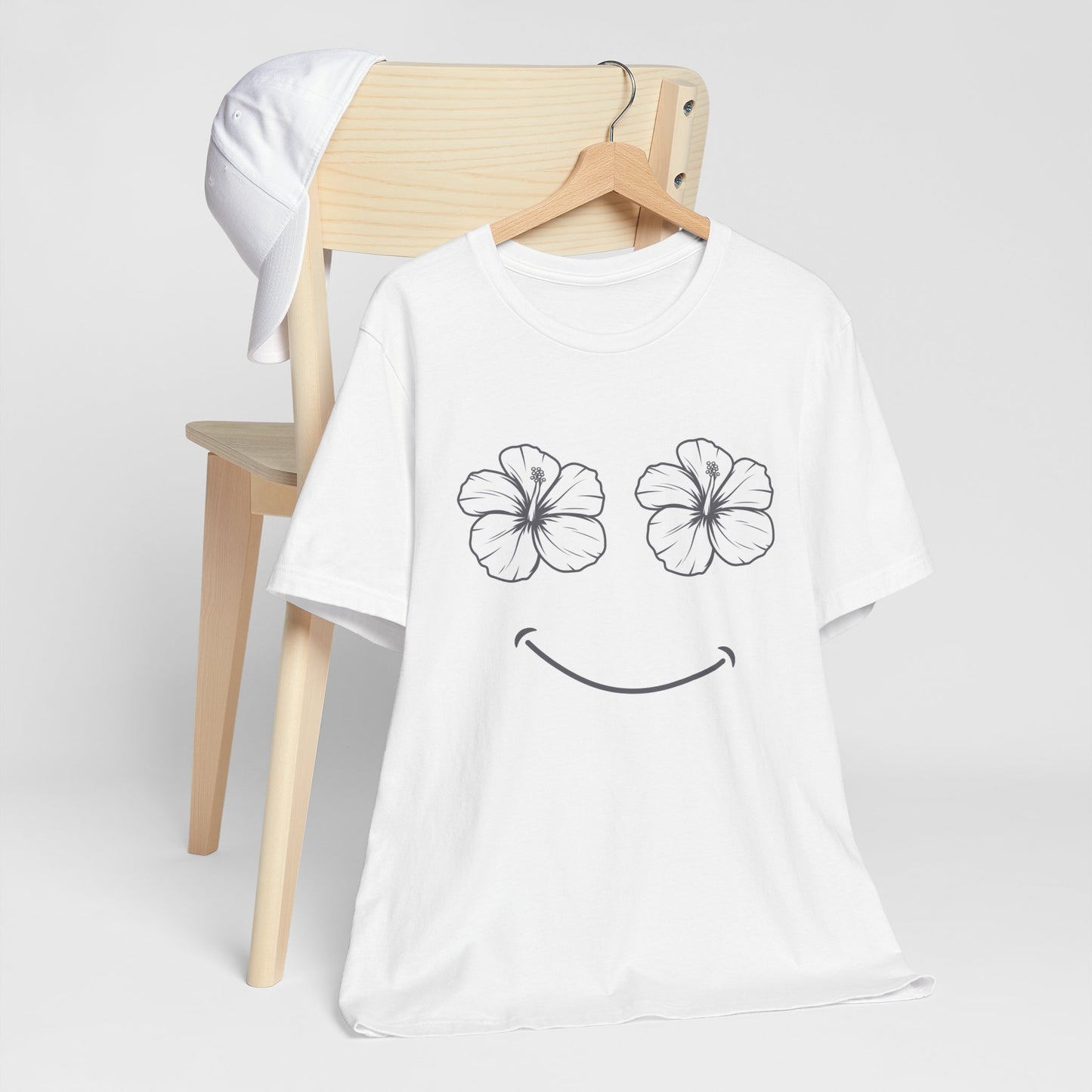 A white short-sleeve t-shirt with a black line-art graphic of two hibiscus flower eyes and a smiley mouth, hanging on a wooden hanger in front of a light wood chair with a white cap draped over it, against a light gray background.