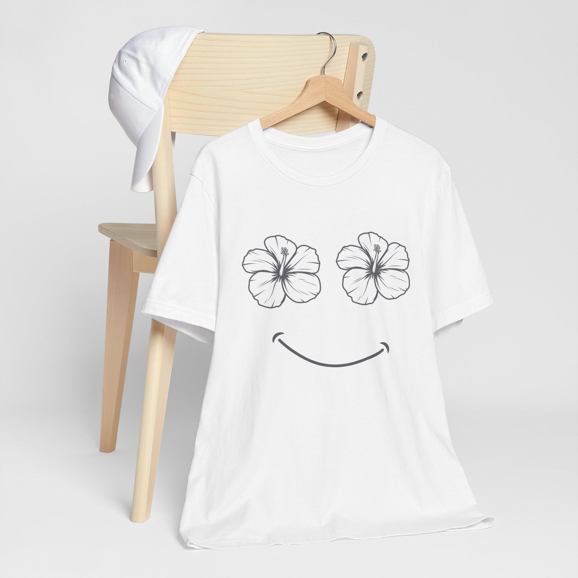 A white short-sleeve t-shirt with a black line-art graphic of two hibiscus flower eyes and a smiley mouth, hanging on a wooden hanger in front of a light wood chair with a white cap draped over it, against a light gray background.