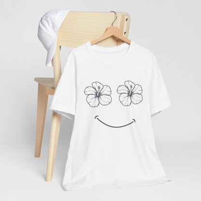 A white short-sleeve t-shirt with a black line-art graphic of two hibiscus flower eyes and a smiley mouth, hanging on a wooden hanger in front of a light wood chair with a white cap draped over it, against a light gray background.