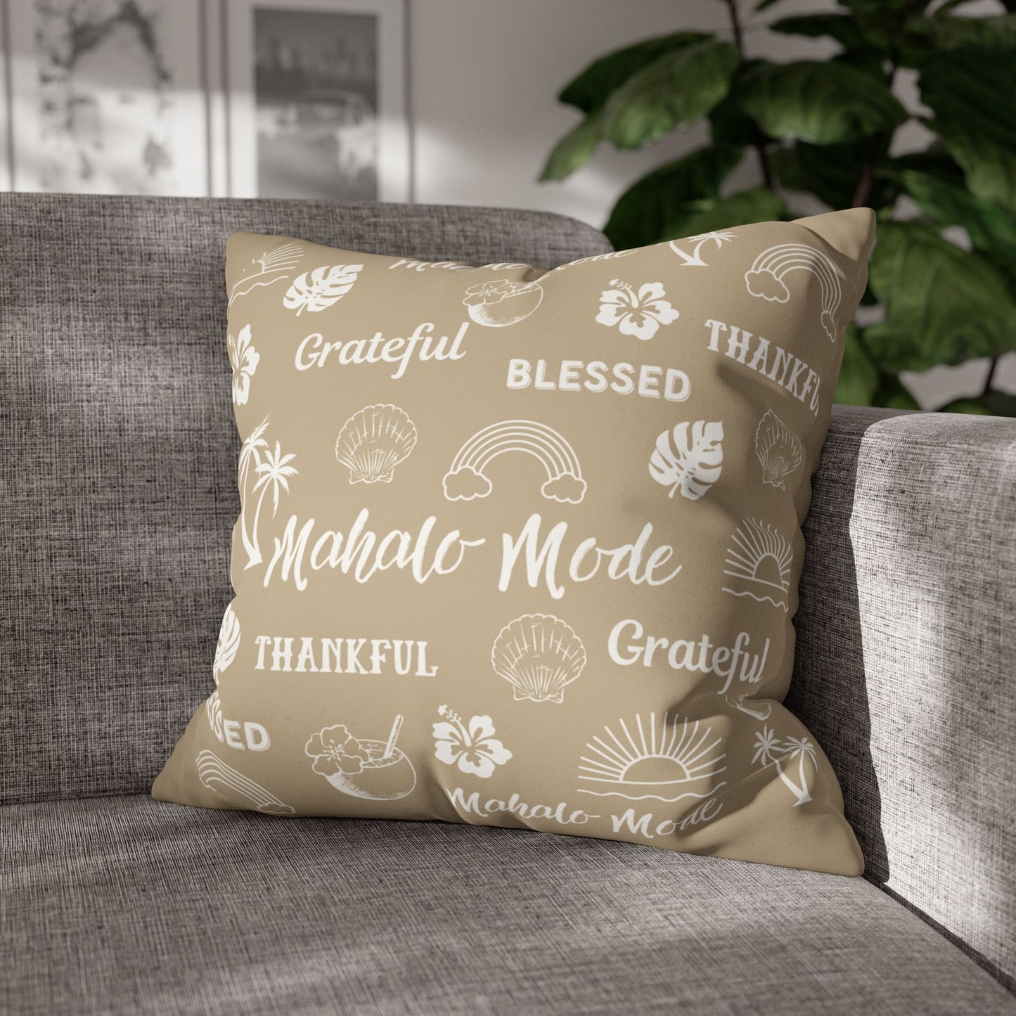 A square throw pillow with a beige background and white tropical-themed text and illustrations, resting on a gray fabric sofa in a living room setting with a green plant in the background.