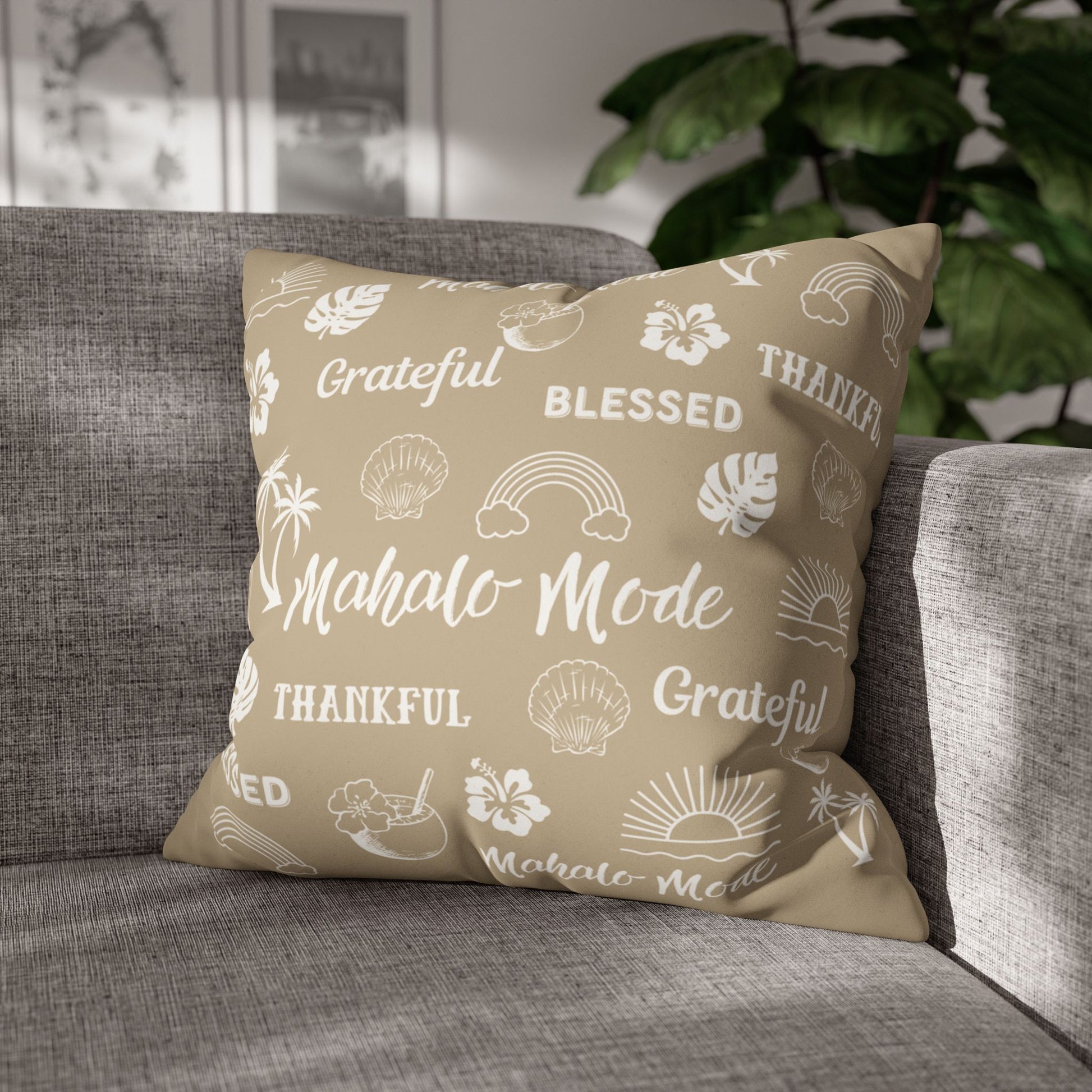 A square throw pillow with a beige background and white tropical-themed text and illustrations, resting on a gray fabric sofa in a living room setting with a green plant in the background.