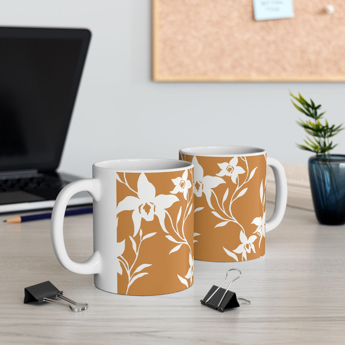 Two white ceramic mugs with a terracotta orange and white floral pattern, displayed on a light wood desk with office props like binder clips, a laptop, and a small plant.