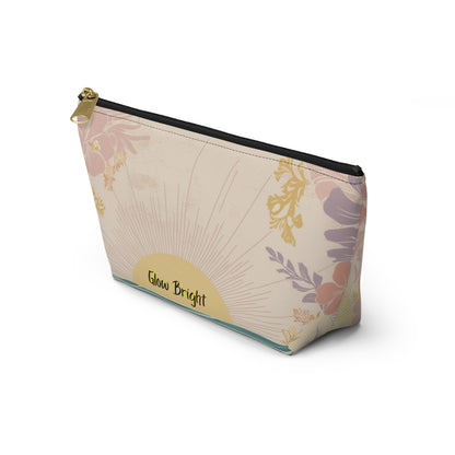 A rectangular zippered pouch with a cream background and a botanical sunburst pattern in muted pink, purple, and gold. A gold oval patch on the front reads 'Glow Bright' in black script.