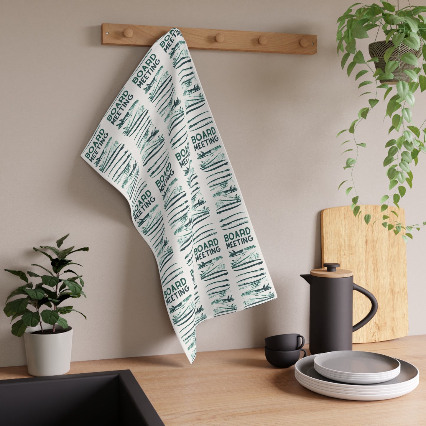 A kitchen towel with a repeating 'BOARD MEETING' text and sketch pattern hanging from a wooden peg on a beige wall in a styled kitchen setting.
