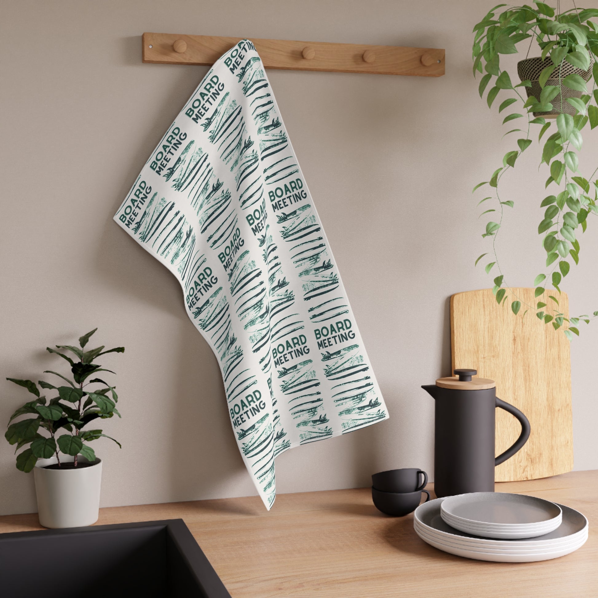 A kitchen towel with a repeating 'BOARD MEETING' text and sketch pattern hanging from a wooden peg on a beige wall in a styled kitchen setting.