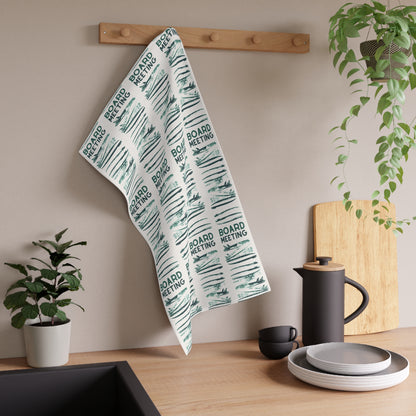 A kitchen towel with a repeating 'BOARD MEETING' text and sketch pattern hanging from a wooden peg on a beige wall in a styled kitchen setting.