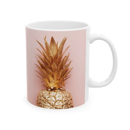 A white ceramic mug featuring a printed graphic of a pineapple with a metallic gold finish against a dusty pink background.